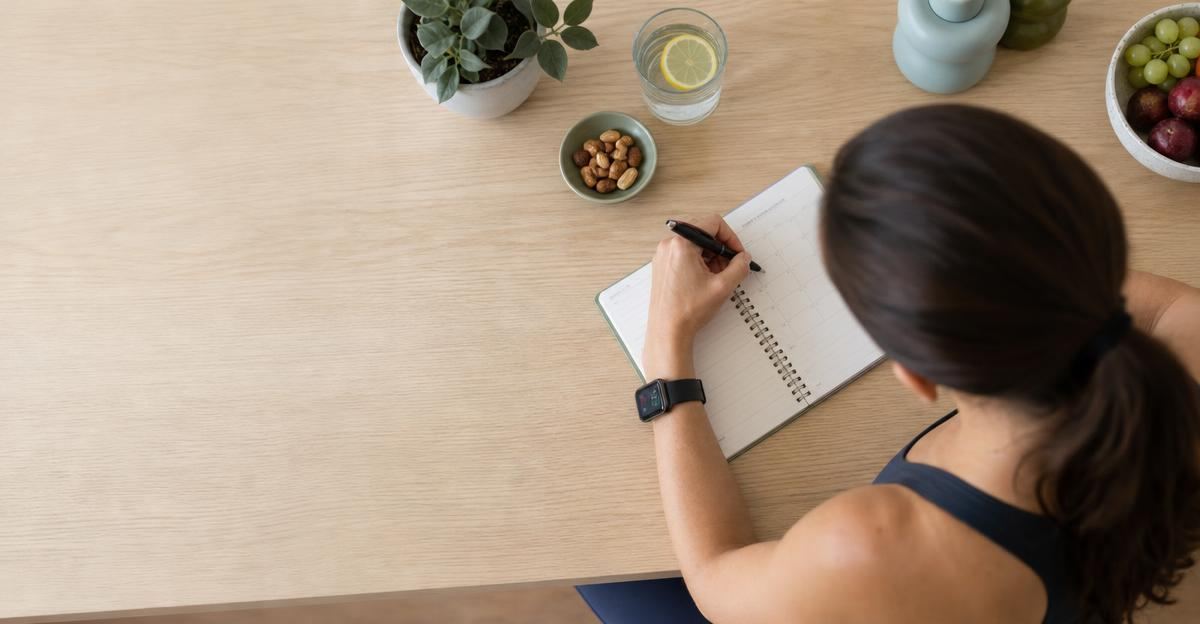 A person sitting at a sunlit wooden desk reviewing health tracking data in a notebook.