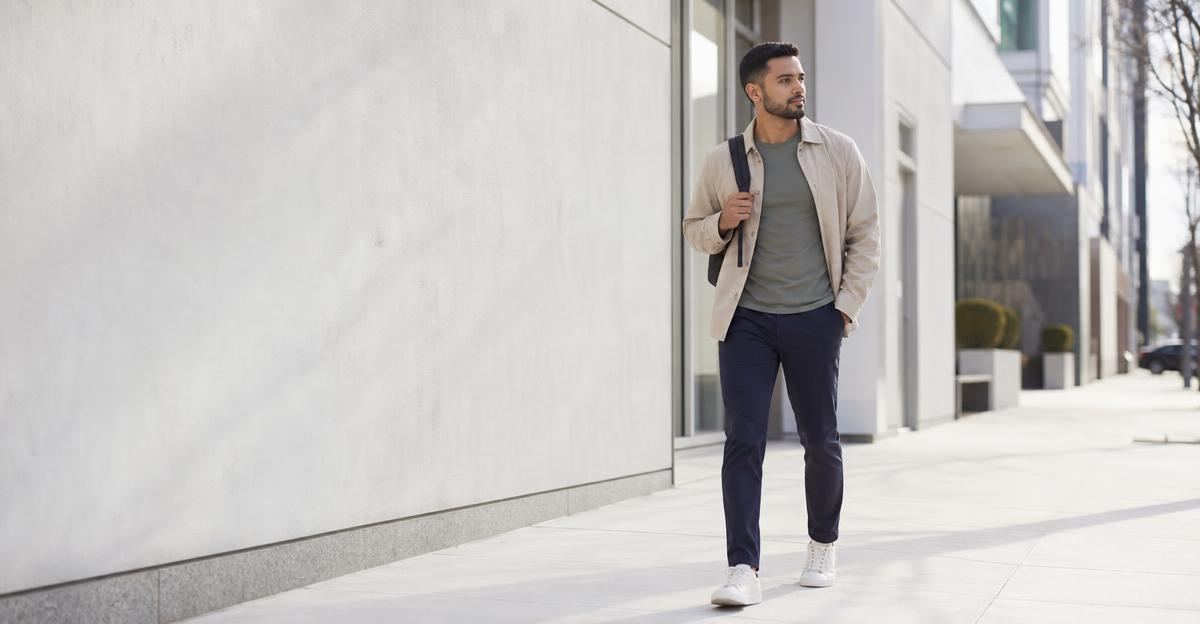 A person in business casual attire walking briskly down a sunlit city sidewalk during a work break.