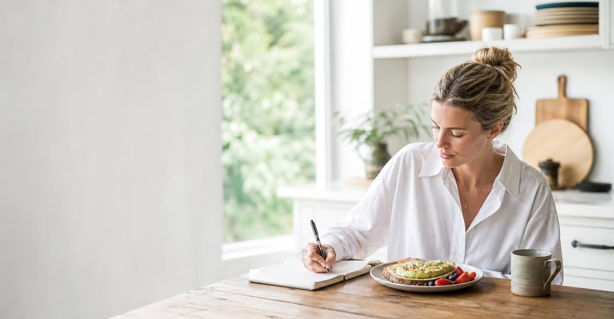 A person sitting at a bright wooden kitchen table, mindfully recording their meal in a notebook.