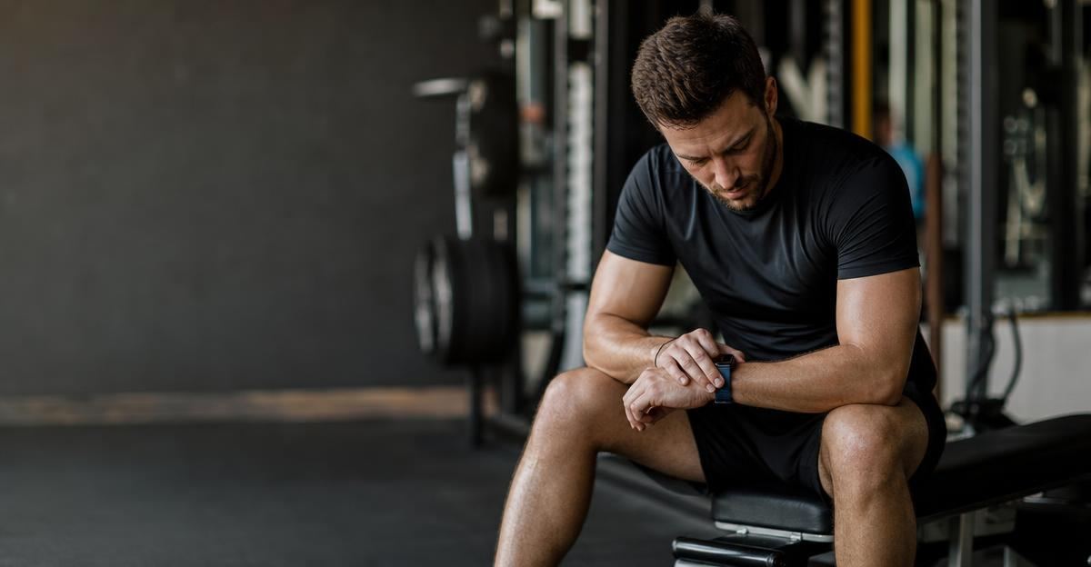 A man in a gym checks his wristwatch while resting between sets during a strength training workout.
