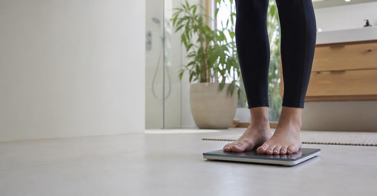 A person wearing gym clothes stands on a digital scale in a brightly lit, modern bathroom.